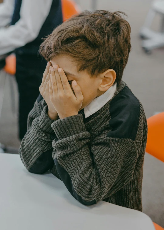 a young boy sitting at a table covering his face, incoherents, in a classroom, anxiety environment, background image, multiple stories