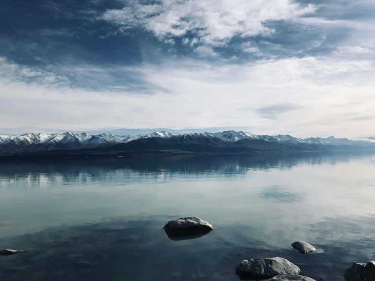 a body of water with rocks and mountains in the background, pexels contest winner, te pae, snow capped mountains, calm clouds, multiple stories