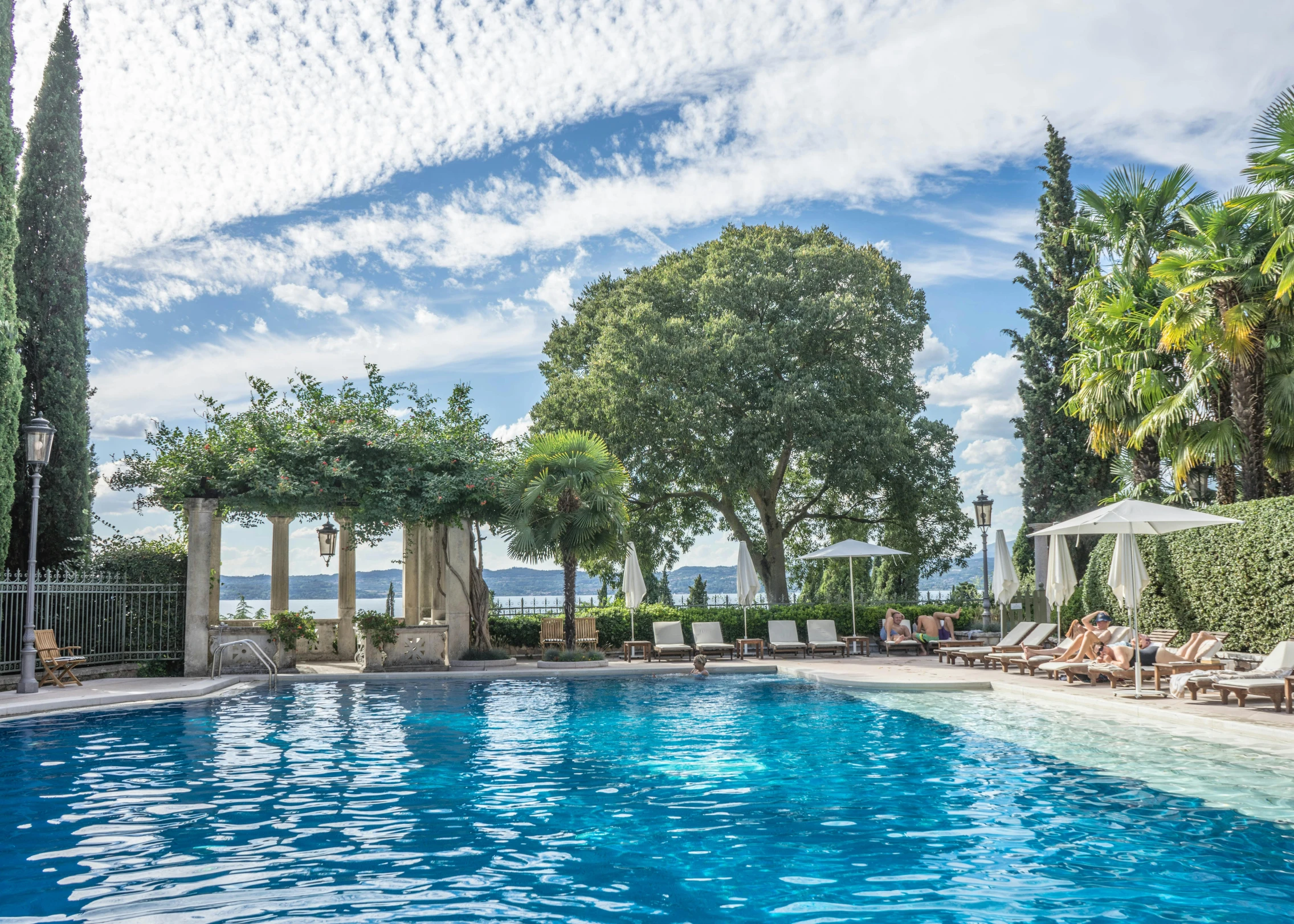 a swimming pool with lounge chairs and umbrellas, pexels contest winner, green grasse trees and river, skies behind, featuring marble fountains, split near the left