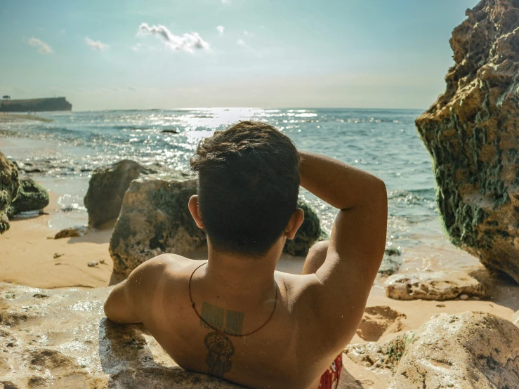 a man sitting on top of a rock next to the ocean, pexels contest winner, happening, deep tan skin, back of head, lgbtq, asian male