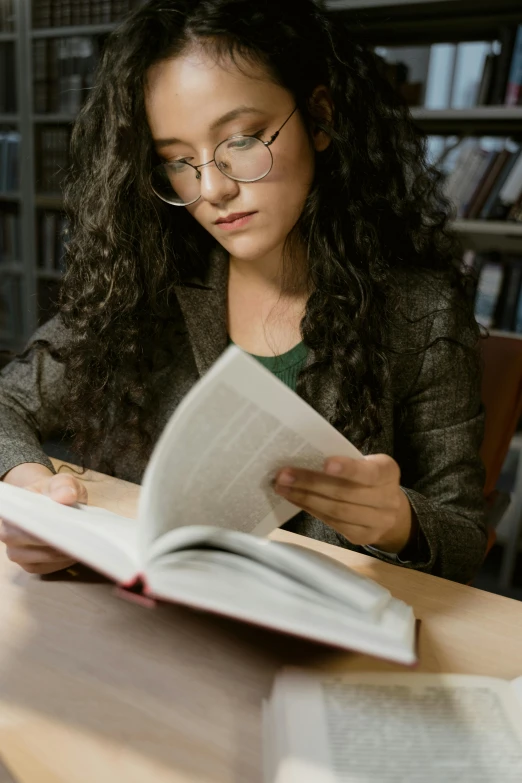 a woman sitting at a table reading a book, wavy long black hair and glasses, college students, on high-quality paper, library books