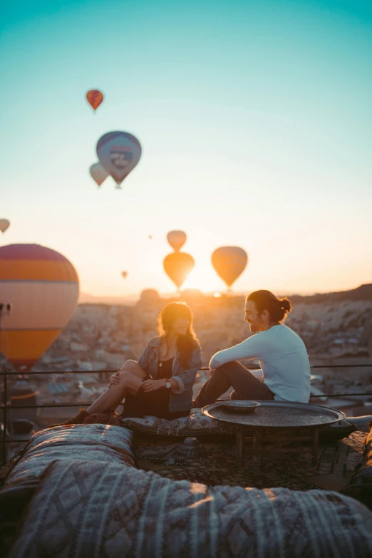 two people sitting on a rock with hot air balloons in the sky, pexels contest winner, happening, sitting on top a table, warm summer nights, staring at you, soft morning light