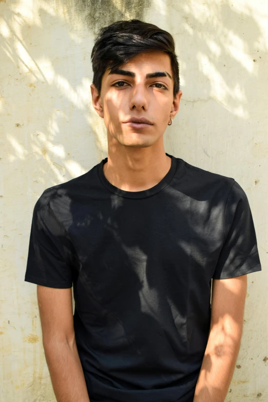 a young man standing in front of a white wall, by Alexis Grimou, pexels contest winner, wearing a black tshirt, middle eastern skin, 1 7 - year - old boy thin face, transgender