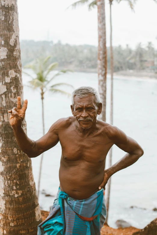 a man standing next to a palm tree in front of a body of water, happening, tribals, the look of an elderly person, waving at the camera, bare chest
