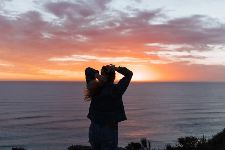 a woman taking a picture of the ocean at sunset, by Jessie Algie, happening, standing on a cliffside, saluting, sunset glow around head, slight overcast