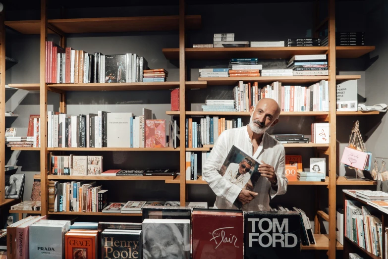 a man standing in front of a bookshelf holding a book, a photo, by Tom Bonson, pexels contest winner, figuration libre, norman foster, sitting on a store shelf, eva elfie, in style of terry richardson