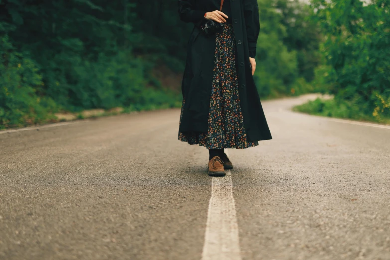 a woman standing in the middle of a road holding an umbrella, trending on pexels, renaissance, long skirt, worn black coat, floral clothes, hilly road