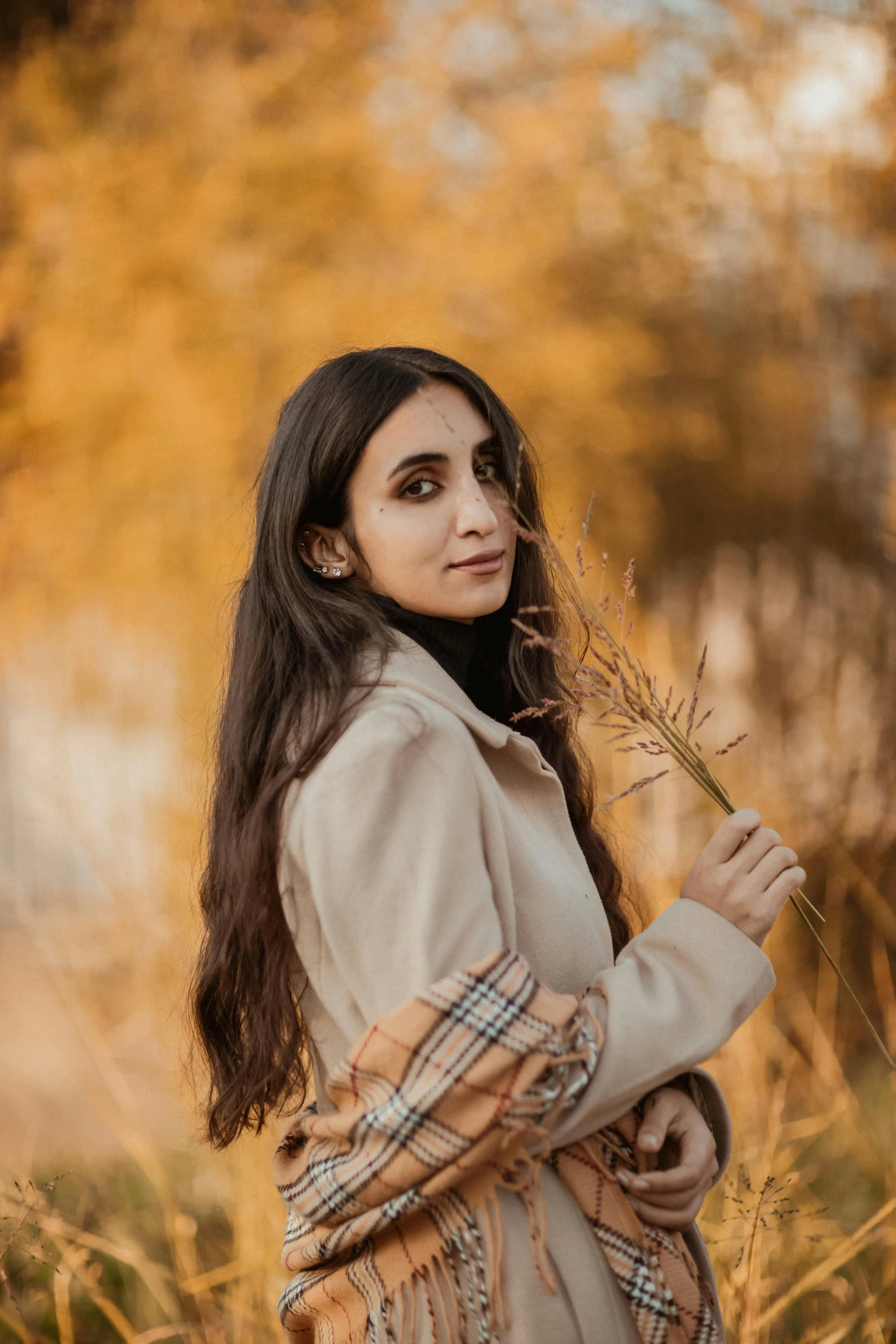 a woman standing in a field of tall grass, a picture, by irakli nadar, trending on pexels, hurufiyya, light brown coat, black hair and large eyes, orange and brown leaves for hair, portait photo profile picture