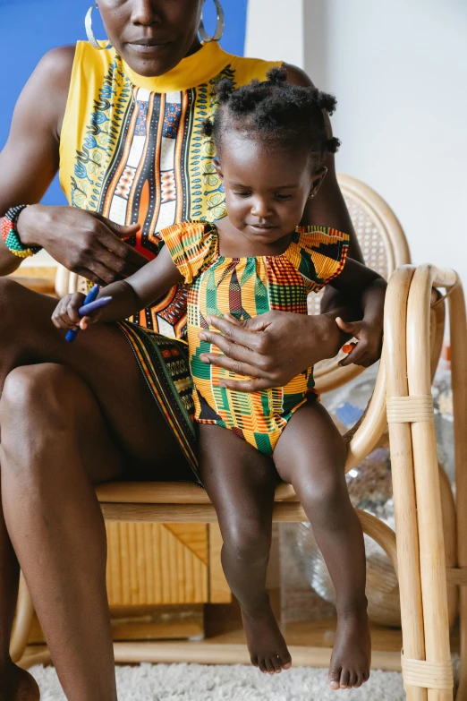 a woman sitting on a chair holding a baby, by Ingrida Kadaka, pexels contest winner, process art, bandage on arms, wearing an african dress, 2 years old, in a gold one piece swimsuit