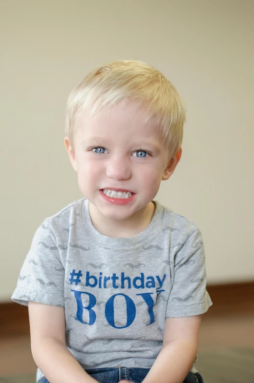 a young boy sitting on the floor wearing a birthday boy t - shirt, blonde hair and blue eyes, thumbnail, all teeth, gray