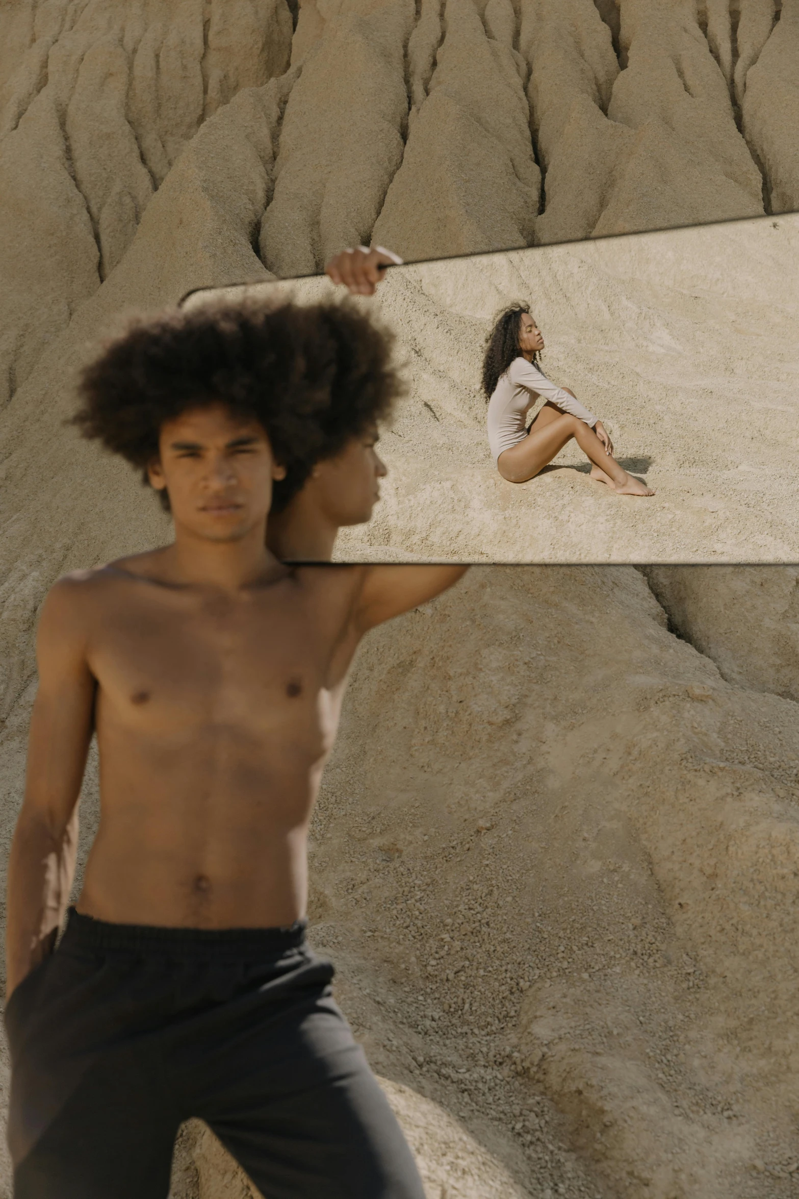 a young man holding a mirror in front of his face, an album cover, by Antoni Pitxot, unsplash, surrealism, his body buried in the sand, long afro hair, two models in the frame, video still