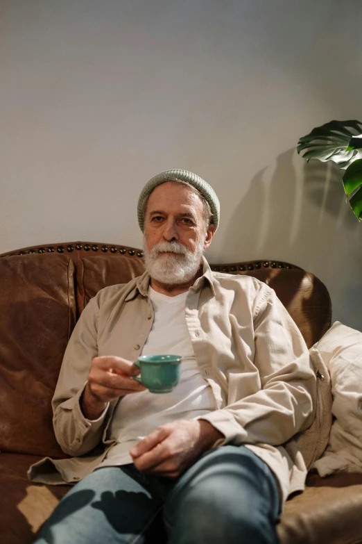 a man sitting on a couch holding a cup, by Leo Michelson, pexels contest winner, short white beard, green corduroy pants, older male, scumbled