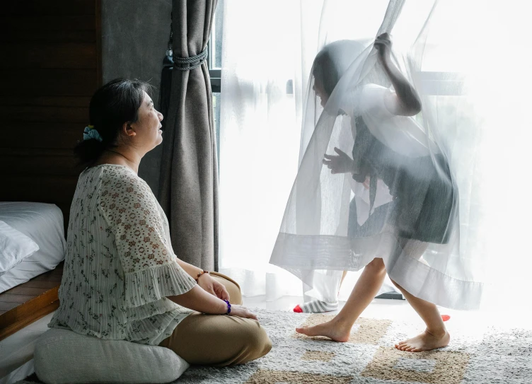 a woman sitting on the floor in front of a window, with a kid, wearing translucent sheet, asian women, kids playing