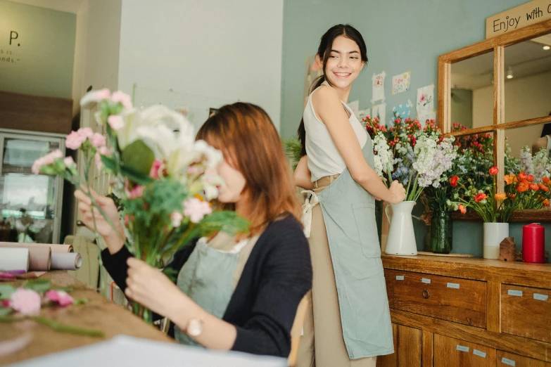 a woman standing in front of a table holding a bunch of flowers, pexels contest winner, arts and crafts movement, woman holding another woman, people at work, white apron, joy ang