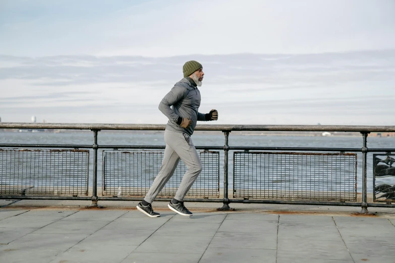 a man riding a skateboard down a sidewalk next to a body of water, inspired by Michael Gustavius Payne, happening, wearing fitness gear, cold weather, grey, people running