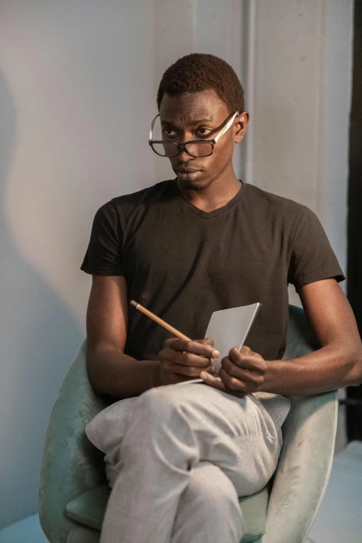 a man sitting in a chair holding a pen and paper, an album cover, inspired by Barthélemy Menn, pexels contest winner, visual art, adut akech, reading glasses, thin young male, holding notebook