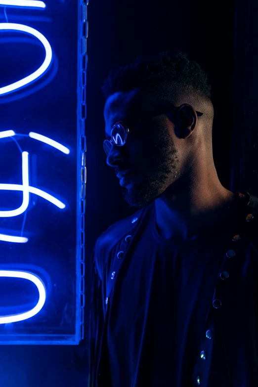 a man standing in front of a neon sign, by Frank Mason, pexels contest winner, afrofuturism, dark blue skin, man with glasses, dark. studio lighting, teddy fresh