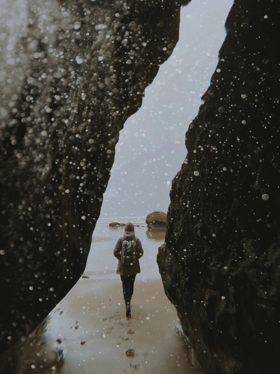 a person walking on a beach between two large rocks, pexels contest winner, snowing outside, droplets on the walls, profile image, looking back at the camera