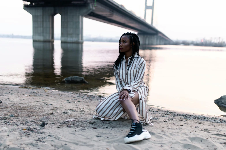 a woman sitting on a beach next to a body of water, a portrait, pexels contest winner, afrofuturism, bridge, female streetwear blogger, portrait of tall, on a riverbank