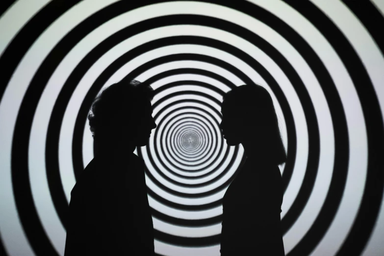 two people standing in front of a black and white spiral background, by Bridget Riley, pexels contest winner, consciousness projection, people's silhouettes close up, symmetrical artwork, exhibit