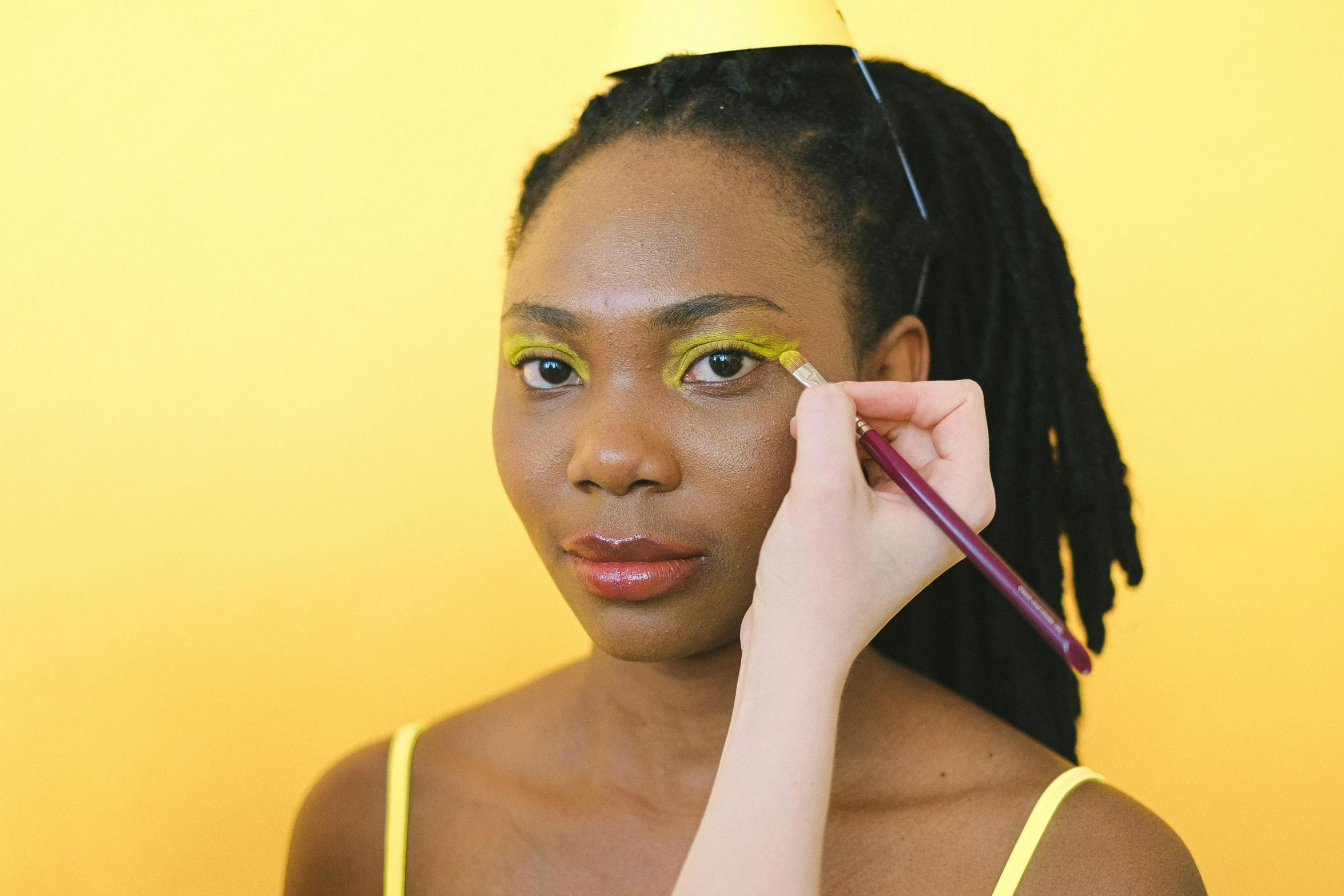 a woman is putting makeup on her face, inspired by Carl Walter Liner, trending on pexels, renaissance, yellow backdrop, complimentary eyeliner, dark skinned, skin painted with green
