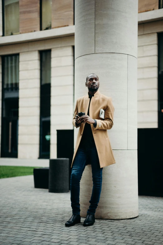 a man leaning against a pillar using a cell phone, inspired by Theo Constanté, pexels contest winner, light brown coat, full body hero, ( ( dark skin ) ), high collar