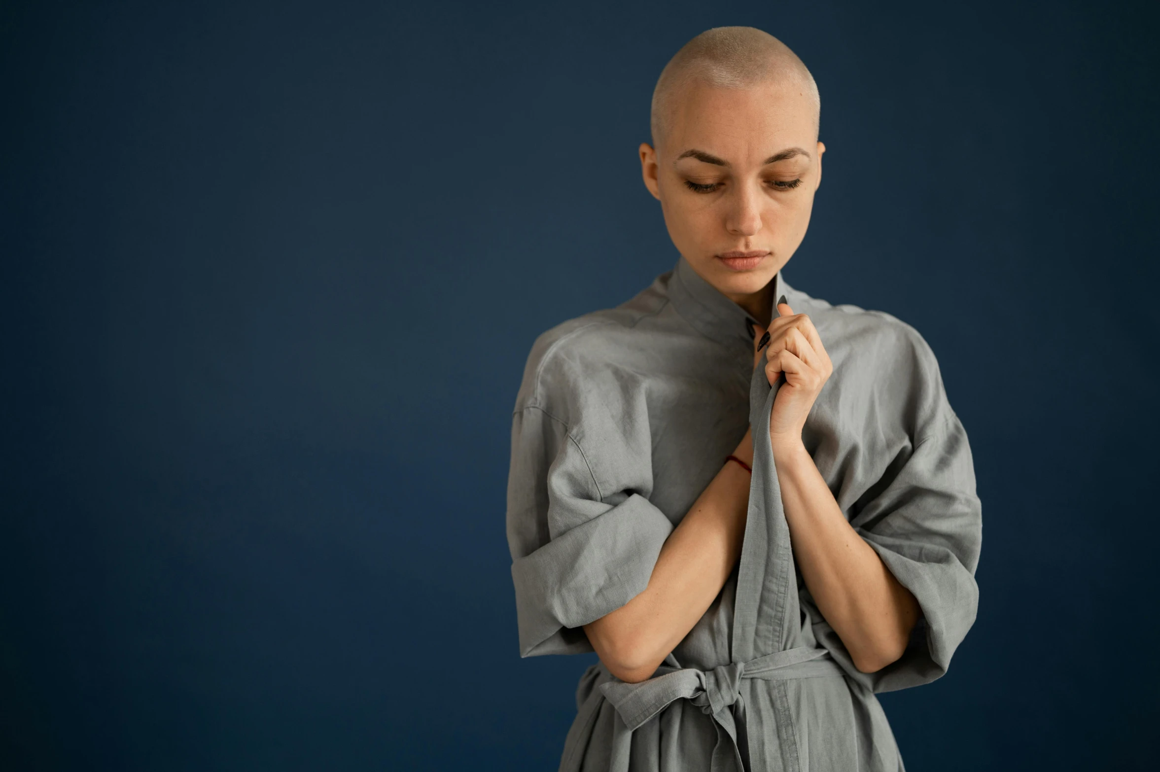 a woman with a shaved head standing in front of a blue background, inspired by Mathieu Le Nain, unsplash, antipodeans, wearing a grey robe, on a gray background, surgical gown and scrubs on, wearing kimono