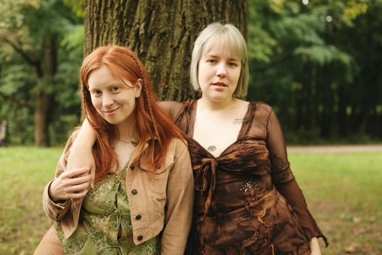 two women standing next to each other near a tree, an album cover, by Grytė Pintukaitė, flickr, maiden with copper hair, green and brown clothes, freckled pale skin, taken in the 2000s