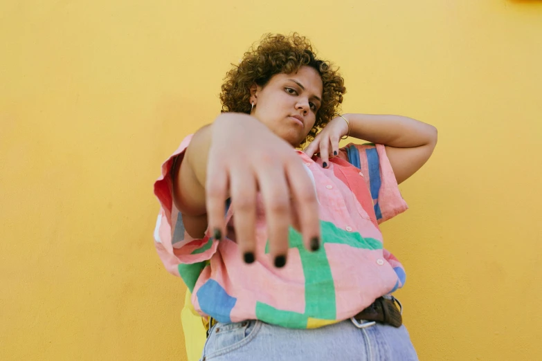 a woman standing in front of a yellow wall, an album cover, by Olivia Peguero, trending on pexels, hyperrealism, gnarled fingers, plus-sized, mixed race, multicoloured