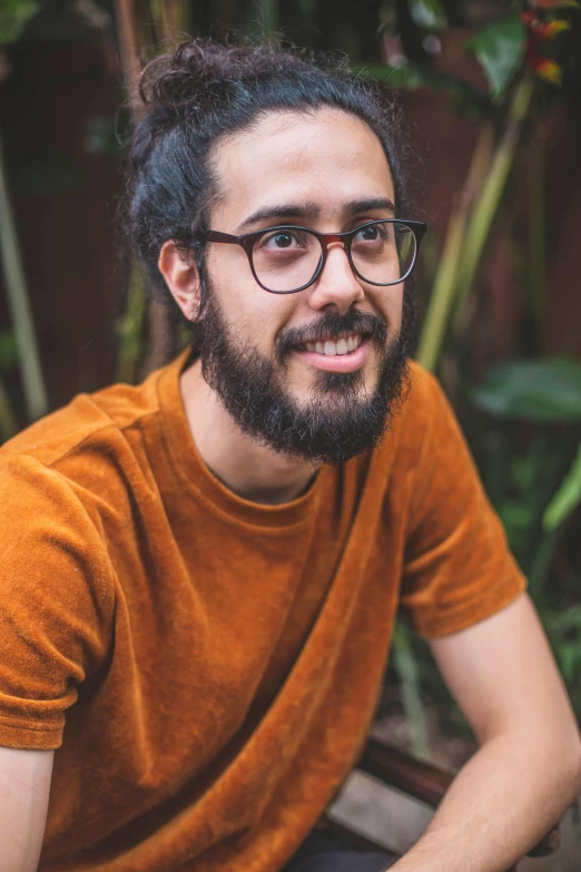 a man with a beard and glasses sitting on a bench, a character portrait, by Alejandro Obregón, pexels contest winner, hurufiyya, in front of a forest background, in front of an orange background, slight nerdy smile, zack de la rocha