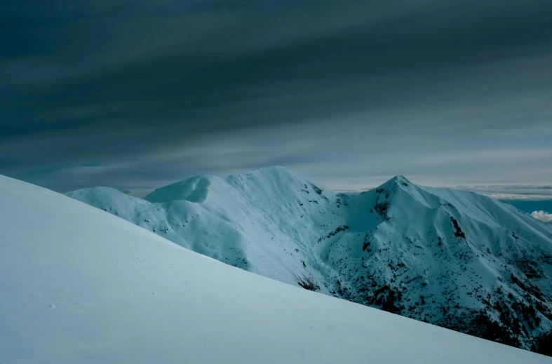 a person standing on top of a snow covered mountain, inspired by Michal Karcz, unsplash contest winner, minimalism, dramatic grey sky, dark winter evening, cold blue colors, irish mountains background