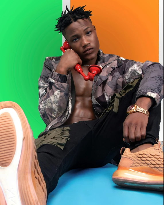 a man sitting on top of a blue table next to a pair of shoes, an album cover, by Cosmo Alexander, trending on pexels, tinyest midriff ever, ( ( ( rainbow ) ) ), posing and flexing, black teenage boy