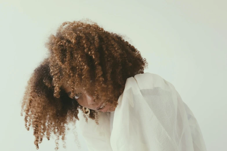 a woman with curly hair wearing a white shirt, by Clifford Ross, trending on pexels, cloak covering face, shy looking down, hair dyed to a hazelnut brown, big red afro