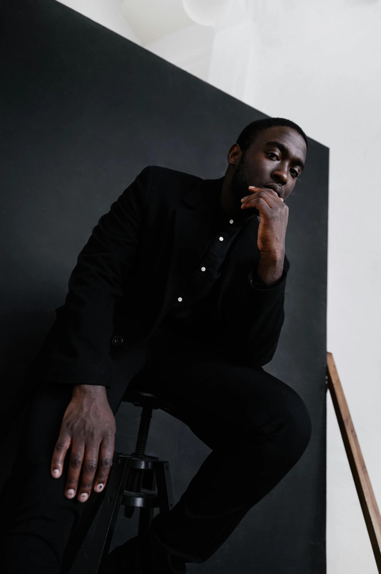 a man sitting on a stool in front of a black wall, inspired by Theo Constanté, jaylen brown, wearing causal black suits, on a pale background, ( ( dark skin ) )