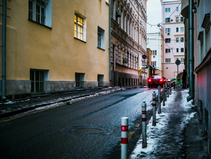 a red car driving down a street next to tall buildings, a photo, pexels contest winner, viennese actionism, cold lighting, cobblestone street, scandinavian, wet asphalt