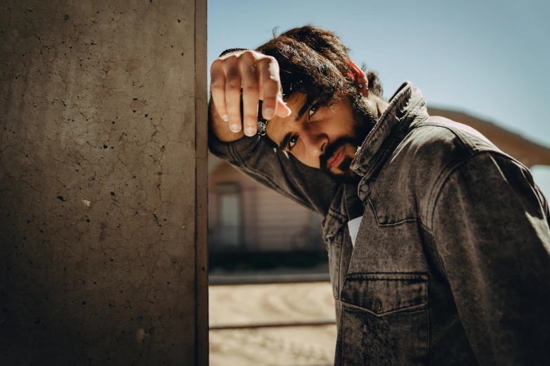 a man in a denim jacket leaning against a wall, pexels contest winner, orelsan, profile image, bearded man, background image