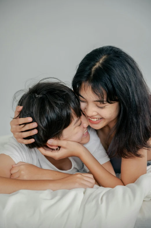 a woman laying on top of a bed next to a child, by Juliette Leong, pexels contest winner, smiling at each other, plain background, male teenager, close face view