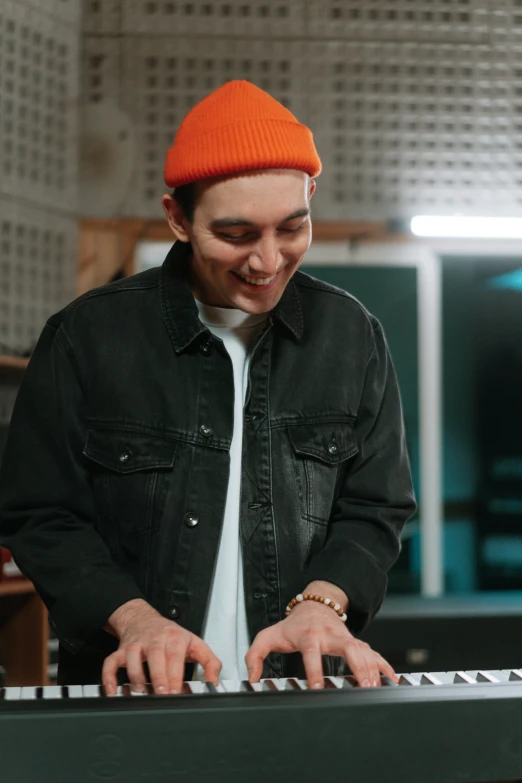 a man that is standing in front of a keyboard, wearing a beanie, orange jacket, in a studio, smiling