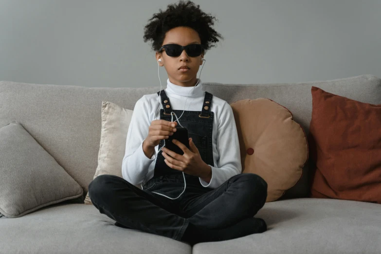 a woman sitting on a couch holding a camera, an album cover, by Adam Marczyński, trending on pexels, afrofuturism, black teenage boy, technological sunglasses, mobile game, little kid