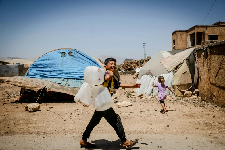 a man walking down a dirt road carrying a bag, a photo, by Youssef Howayek, hurufiyya, makeshift houses, water to waste, thumbnail, camps in the background