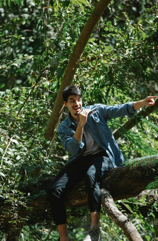 a man sitting on top of a tree branch, happening, pointing at the camera, rex orange county, avan jogia angel, enjoying a stroll in the forest