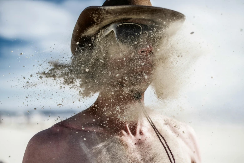 a man standing on top of a sandy beach, inspired by Russell Drysdale, pexels contest winner, process art, hat covering eyes, floating dust particles, portrait of a rugged ranger, clean face and body skin