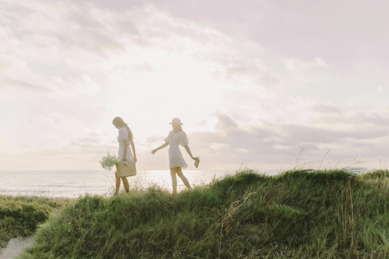 a couple of people standing on top of a grass covered hill, by Jessie Algie, unsplash, walking on the beach, two girls, carrying flowers, pale light