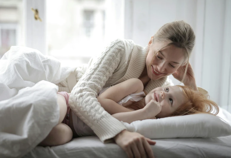 a woman laying on top of a bed next to a little girl, smiling at each other, soft and warm, commercially ready, white