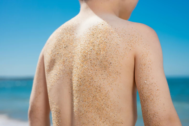 a boy standing on a beach covered in sand, trending on pexels, showing her shoulder from back, sand particles, sunbathed skin, spangle