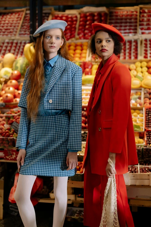 two women standing next to each other in front of a fruit stand, an album cover, by Anita Malfatti, trending on pexels, renaissance, red tailcoat, blue coat, chequered cape, editorial model