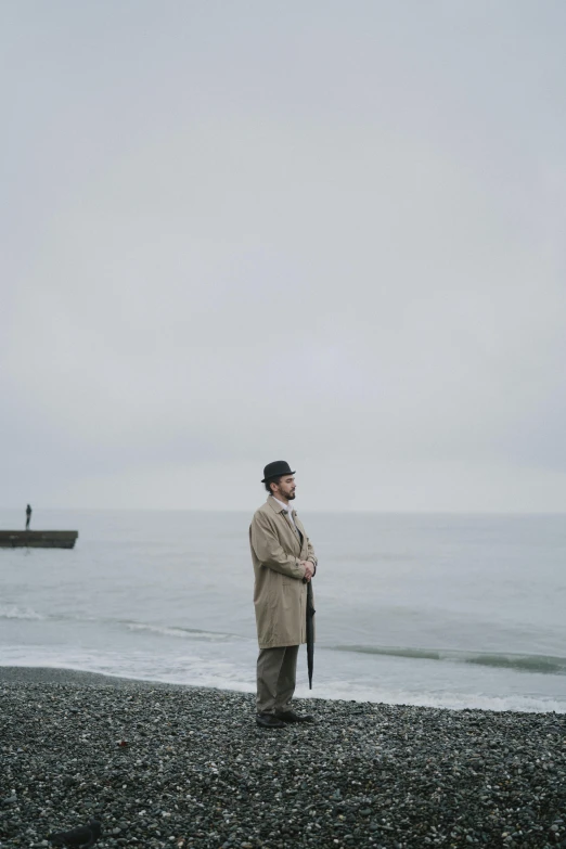 a man standing on top of a beach next to the ocean, trench coat, grey skies, a suited man in a hat, [ cinematic