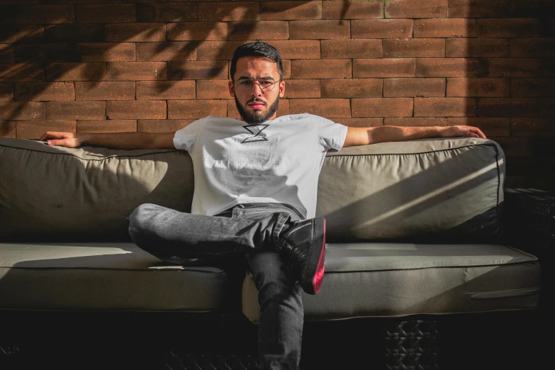 a man sitting on a couch in front of a brick wall, inspired by Nabil Kanso, pexels contest winner, les nabis, wearing pants and a t-shirt, avatar image, profile image, attractive photo