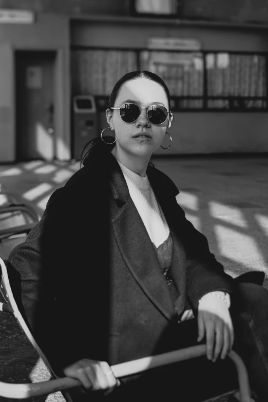 a black and white photo of a woman sitting on a bench, a black and white photo, inspired by Fei Danxu, unsplash, bauhaus, round sunglasses, wearing a turtleneck and jacket, portrait sophie mudd, trench coat and suit