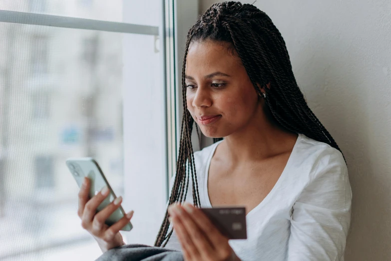 a woman sitting on a window sill looking at her cell phone, pexels contest winner, renaissance, holding an ace card, african american young woman, at checkout, woman with braided brown hair